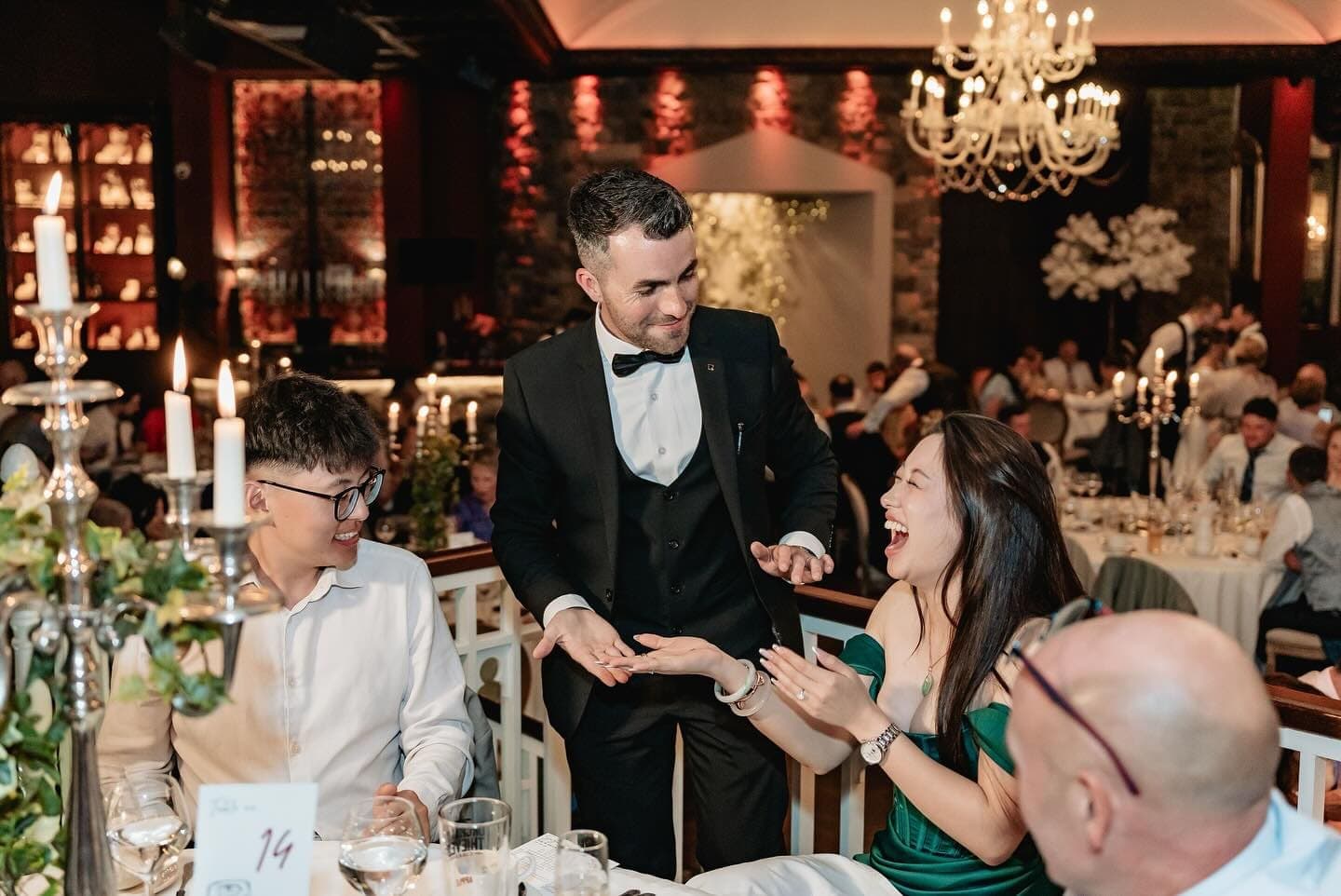At Alex and Suzanne's wedding, the guest in green laughs out loud at Jamie's hands as he leans in at the table