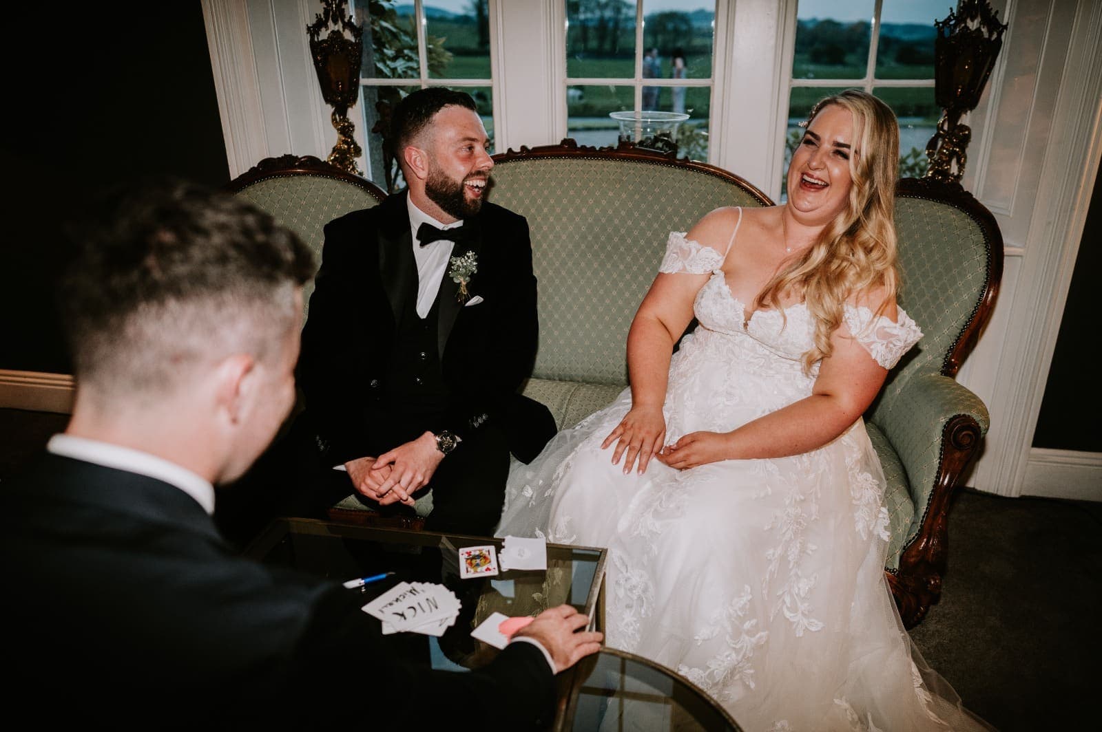 At Janice and Oisin's wedding at Clonabreany House, Janice laughs on a green sofa as Oisin smiles and Jamie deals cards