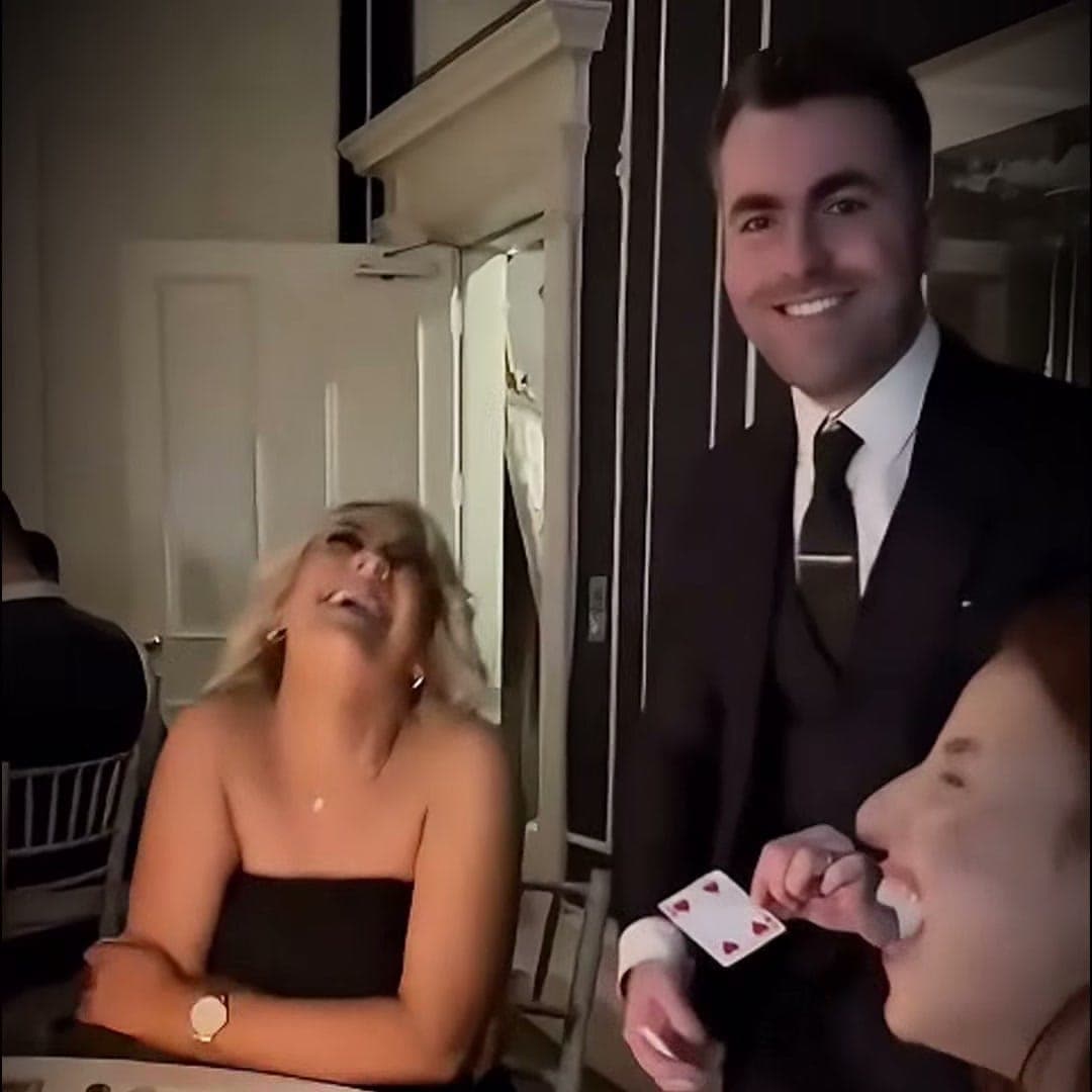 A guest in a black dress laughs with her head back as Jamie smiles and holds up the 4 of Hearts