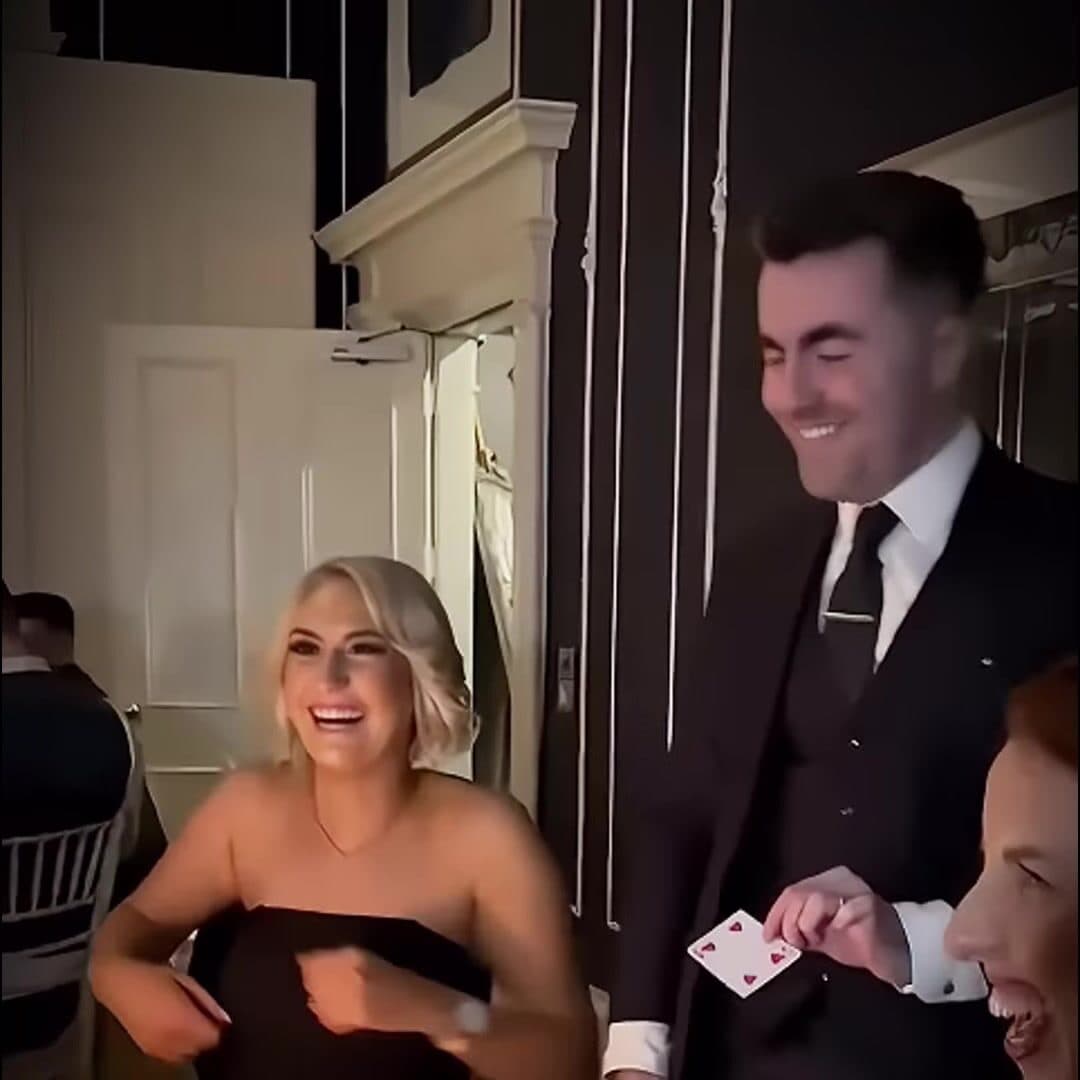 Jamie smiles and holds up the 4 of Hearts while two guests at the table laugh