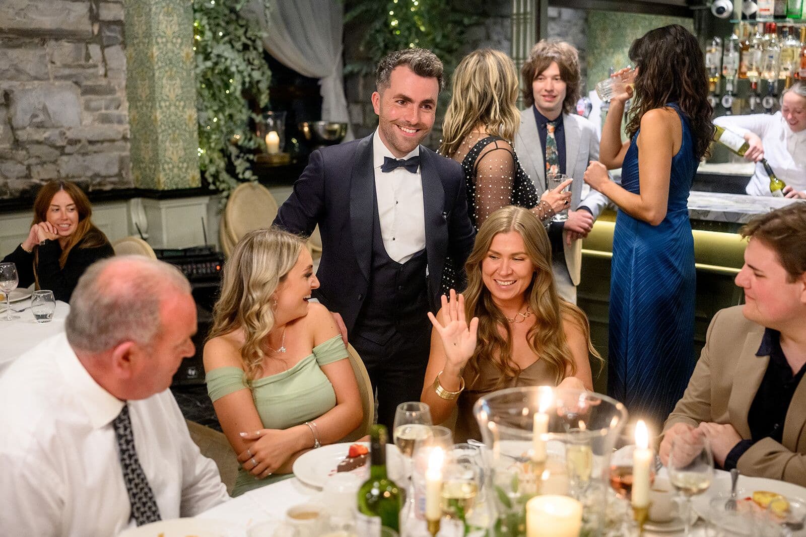 Jamie stands behind a table of smiling guests, one of them waving