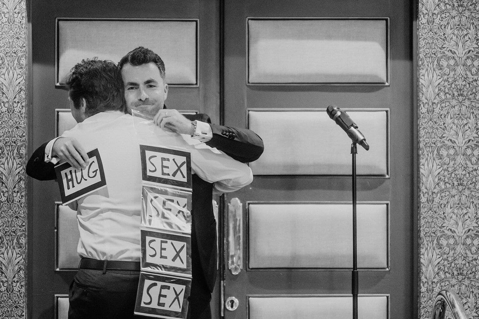 Black and white: Jamie hugs a guest who has cards on his back, holding a card that reads HUG