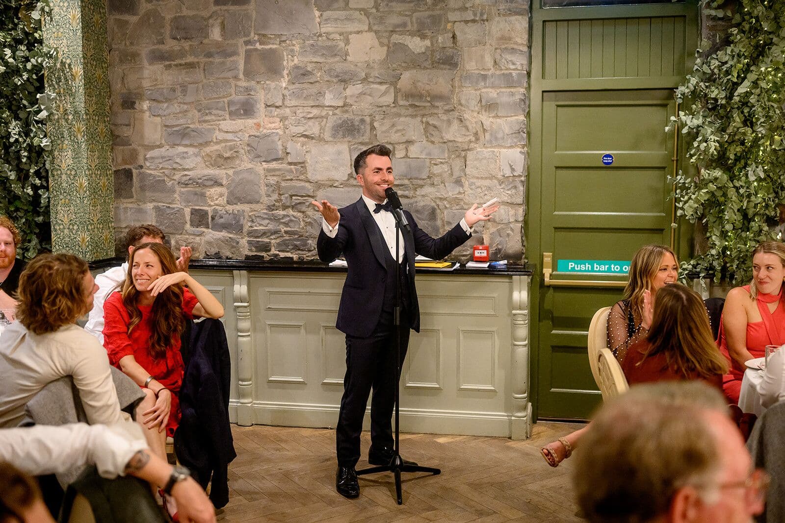Jamie, on the mic with arms wide, hosts the stone-walled reception room