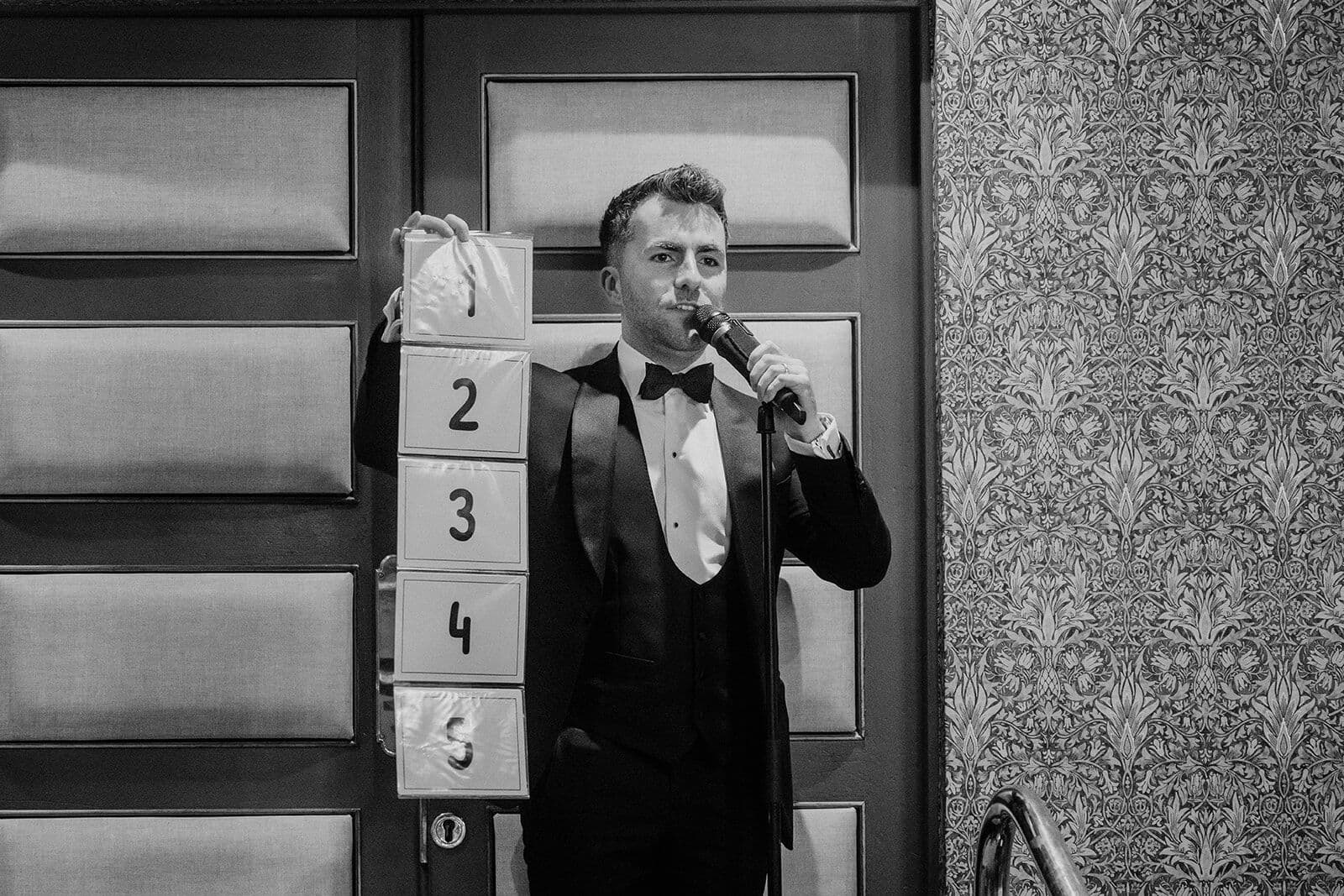 Black and white: Jamie on the mic holds up a strip of cards numbered 1 to 5