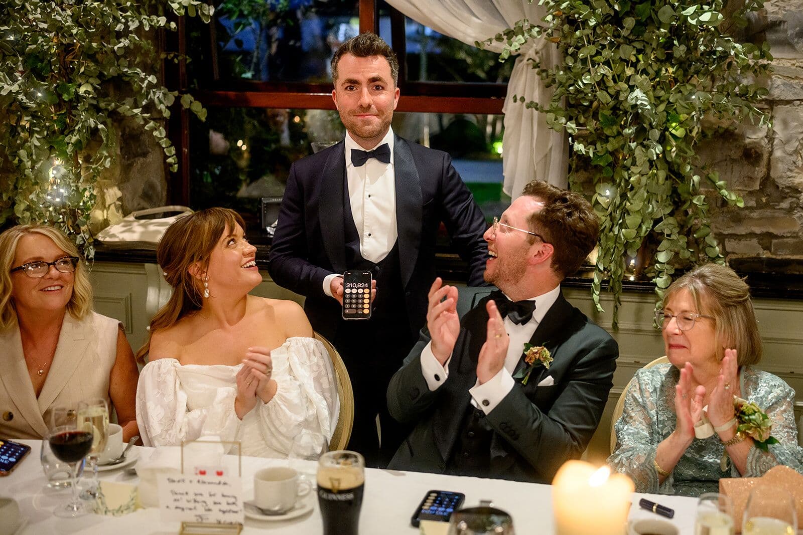 Alexandra and Travis clap at the top table at The Station House Hotel as Jamie holds up a phone behind them