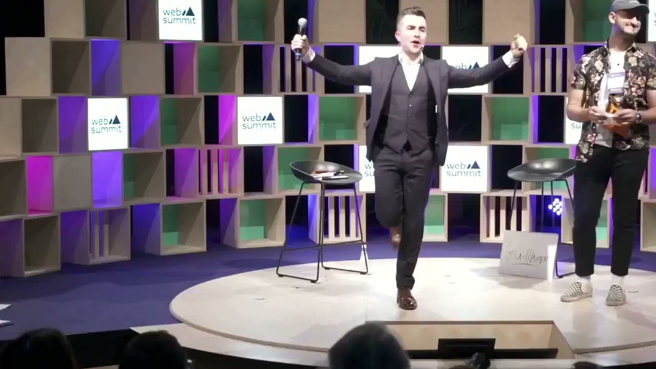 Jamie, microphone in hand, throws his arms wide on a conference stage beside a volunteer, the audience in front