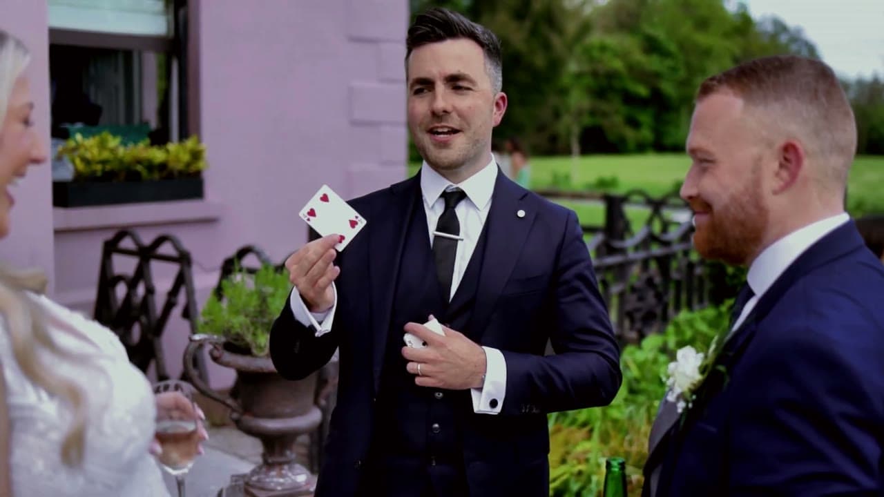 Jamie smiles as he holds up a playing card to a smiling groom and a laughing bride at a garden wedding