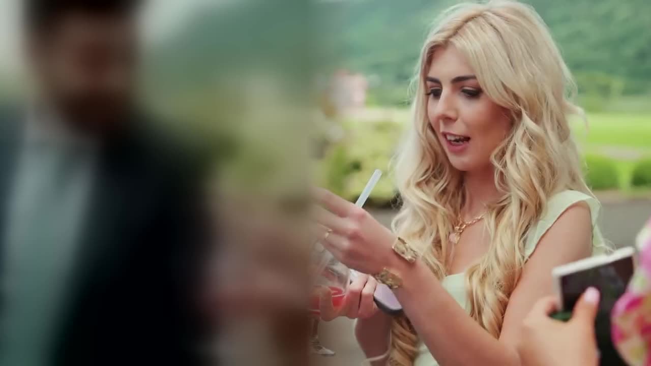 A woman with long blonde hair gasps at a card trick on a lawn, green hills behind her