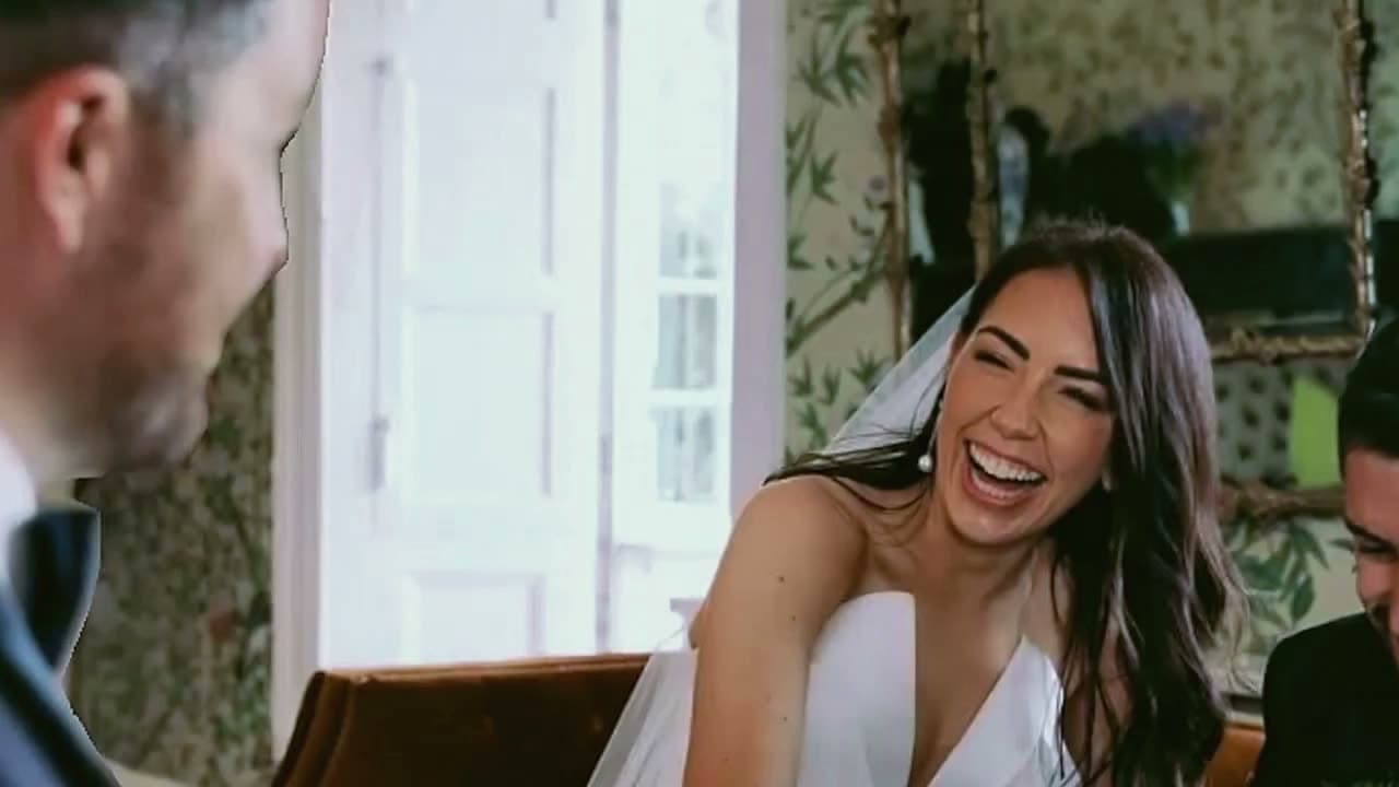 A bride in a white dress laughs at a trick beside a guest in a sitting room
