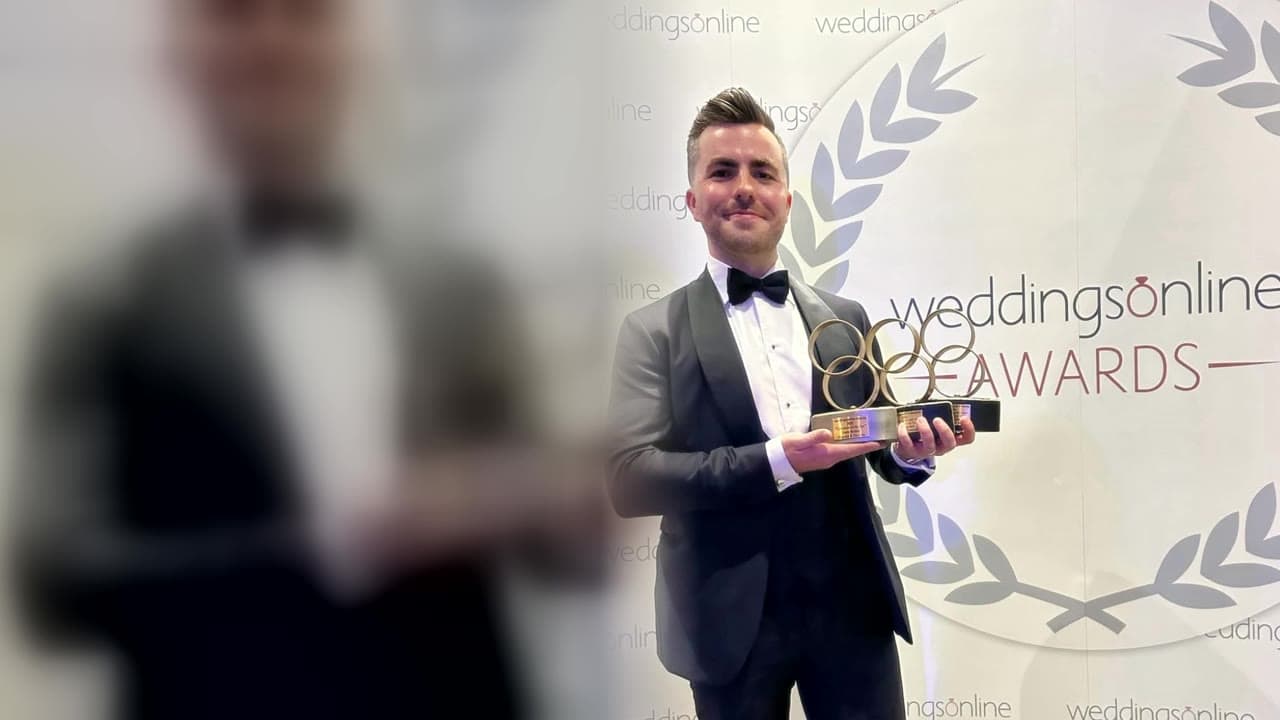 Jamie in a tuxedo smiles as he holds three weddingsonline awards in front of the weddingsonline Awards wall