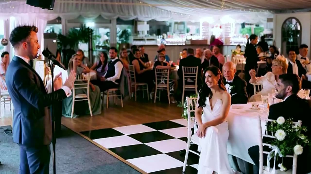 Jamie speaks into a microphone at a marquee wedding as the bride smiles up at him from the top table