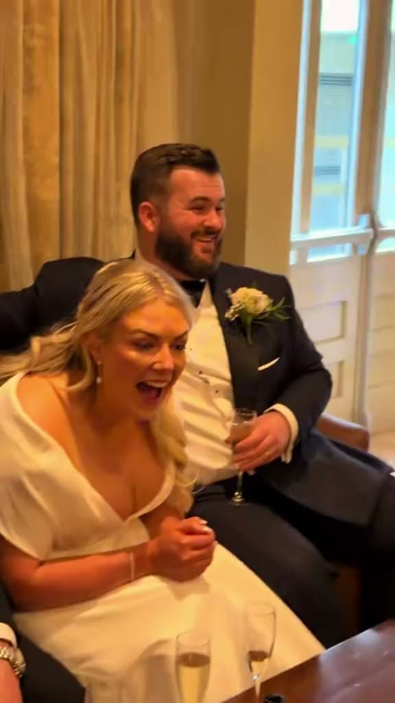 A laughing bride with her mouth wide open sits on a sofa beside the groom, who grins at something out of frame